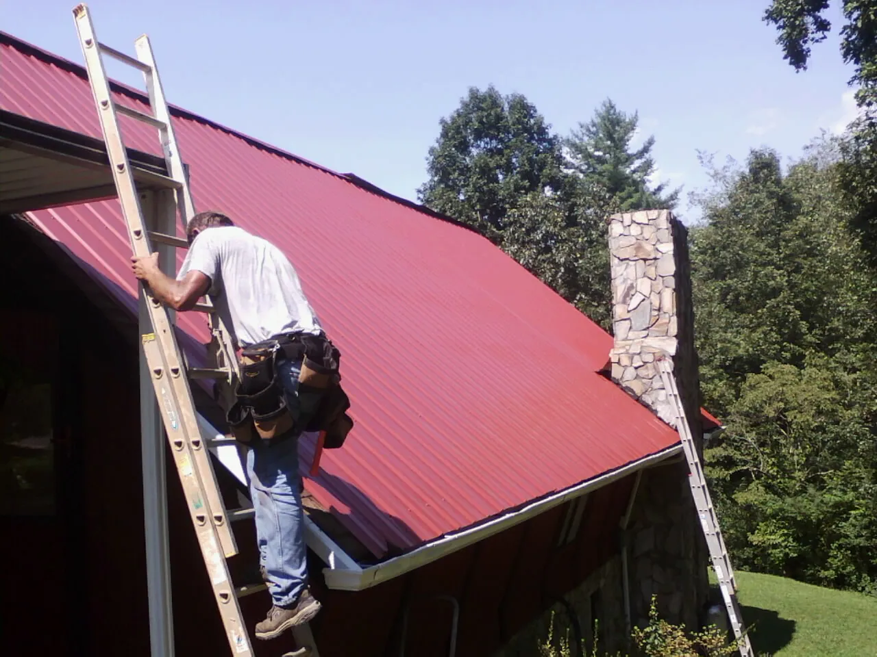 Roofer on a ladder beside a red metal roof and stone chimney — Soffit & Fascia Repair by Roofing Brothers in Highspire